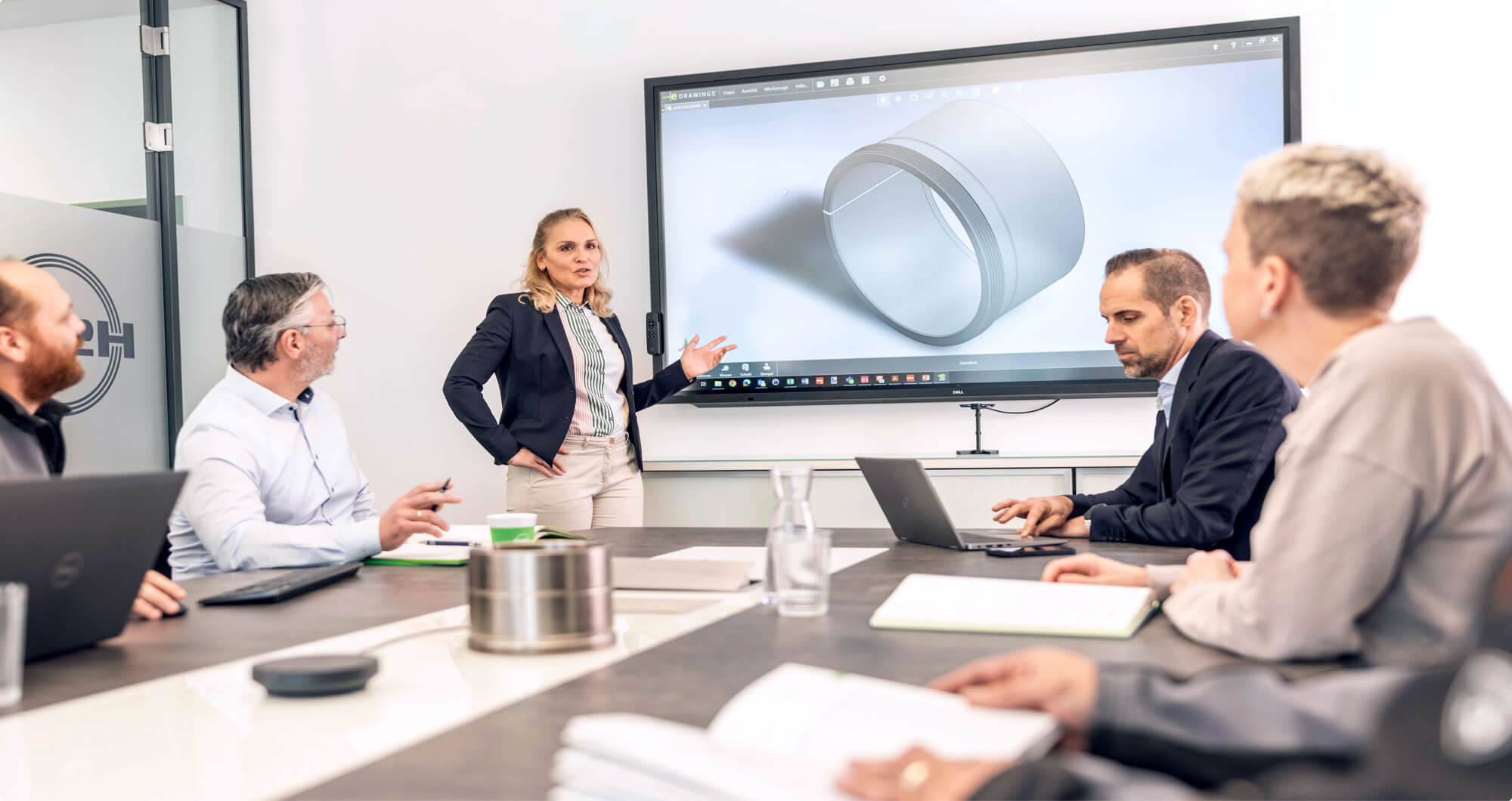 Person präsentiert in einem Meeting eine technische 3D-Zeichnung auf einem großen Bildschirm, während mehrere Kollegen aufmerksam zuhören und Notizen machen.